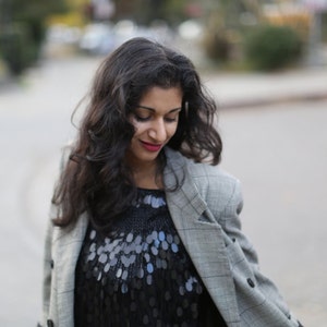 May include: A woman with long dark hair is wearing a gray blazer over a black sequined top. She is looking down at something off camera.