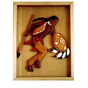 May include: A wooden carving of a Native American warrior in a brown and orange outfit, holding a spear and a shield with feathers. The carving is framed in a light wood frame.