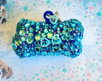 May include: A blue and green jeweled dog bone charm with a gold chain attached. The charm is covered in rhinestones and has the word "Dog" spelled out in white beads.