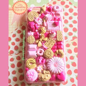 May include: A pink phone case decorated with various pink, white, and gold candies and sweets. The case is covered in a variety of treats, including cookies, biscuits, chocolate bars, and sprinkles. The case is surrounded by a pink and white polka dot background.