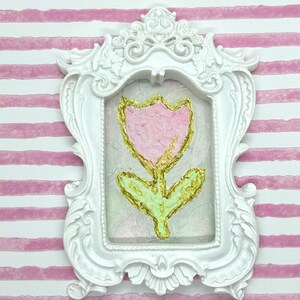 May include: A white ornate picture frame with a pink tulip painting. The tulip has gold accents and green leaves. The background is a light pink and the frame is set against a pink and white striped background.