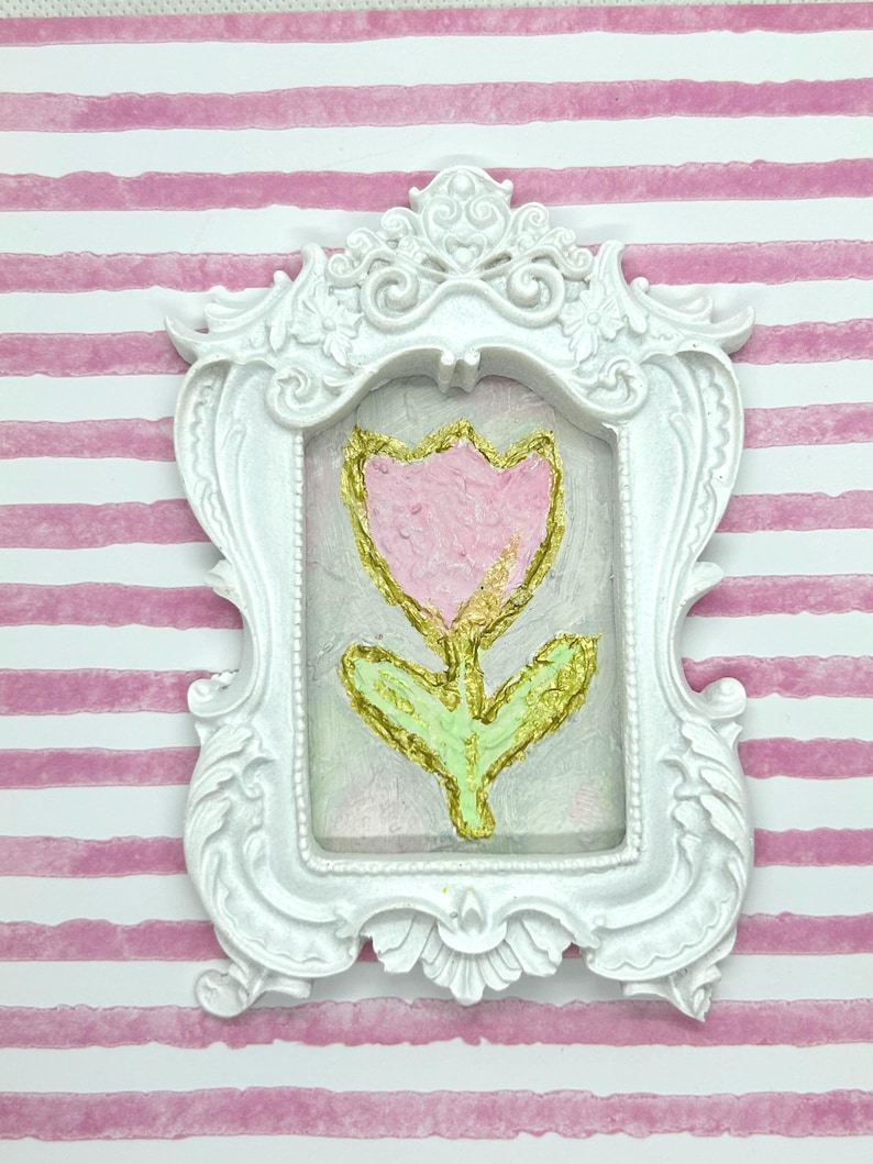 May include: A white ornate picture frame with a pink tulip painting. The tulip has gold accents and green leaves. The background is a light pink and the frame is set against a pink and white striped background.