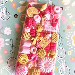 May include: A pink phone case decorated with a variety of colorful 3D food-shaped embellishments, including cookies, candy, and chocolate. The case features the words "Bonjour" and "Biscuit" in pink lettering.
