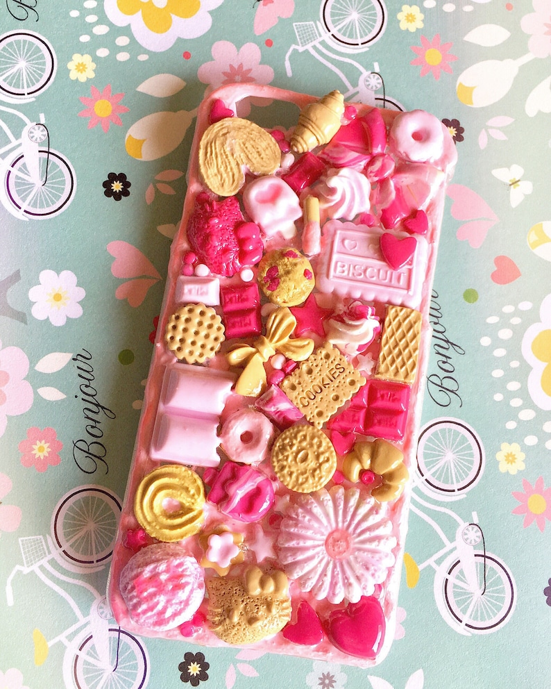 May include: A pink phone case decorated with a variety of colorful 3D food-shaped embellishments, including cookies, candy, and chocolate. The case features the words "Bonjour" and "Biscuit" in pink lettering.