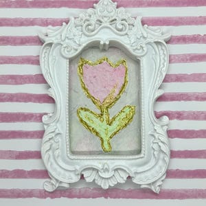 May include: A white ornate frame with a pink and gold flower painting. The flower is a tulip shape with a pink petal and gold accents. The background is a light, textured surface.