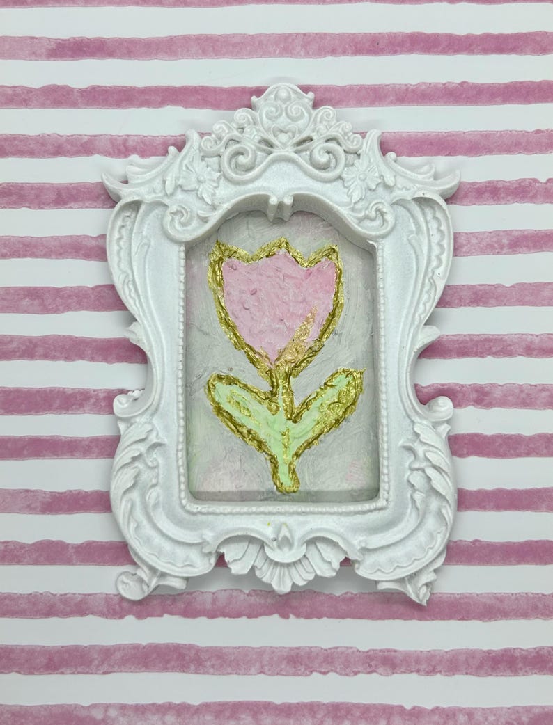May include: A white ornate frame with a pink and gold flower painting. The flower is a tulip shape with a pink petal and gold accents. The background is a light, textured surface.