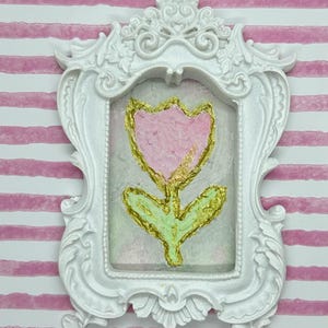 May include: A white ornate picture frame with a pink and gold tulip painting. The tulip has a pink petal and a gold outline. The stem and leaves are green with gold accents. The background is a light green and pink.