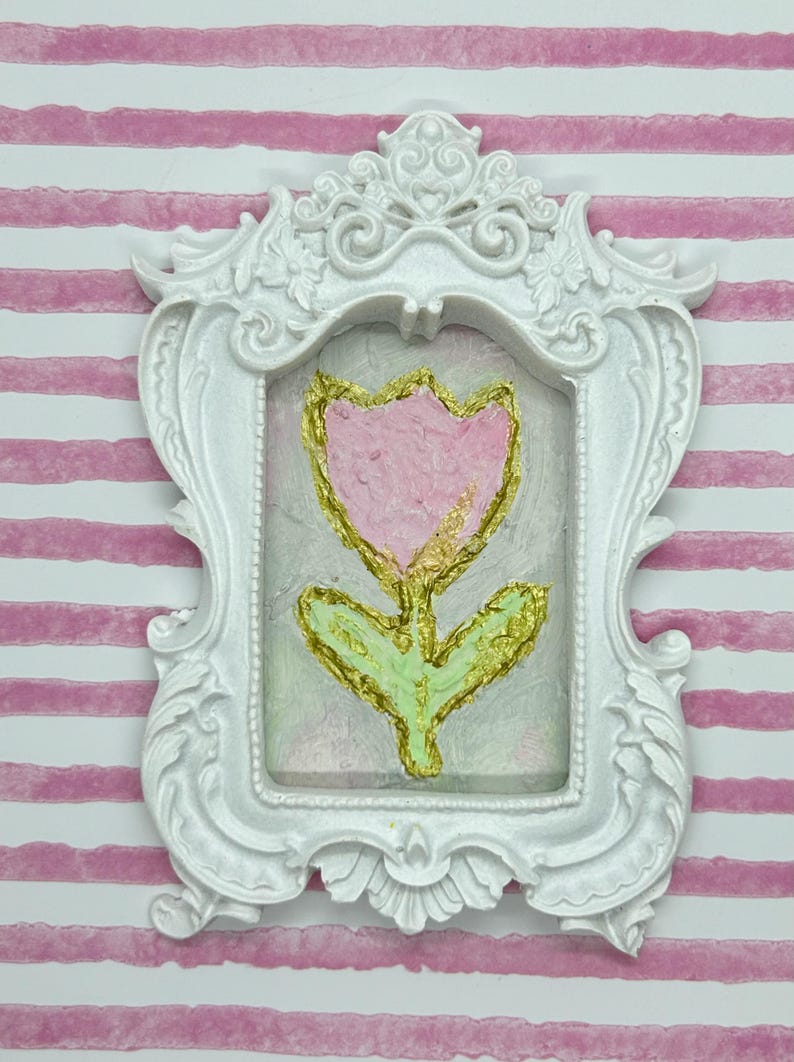 May include: A white ornate picture frame with a pink and gold tulip painting. The tulip has a pink petal and a gold outline. The stem and leaves are green with gold accents. The background is a light green and pink.