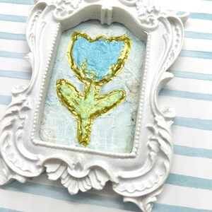 May include: A small, white, ornate picture frame with a painted tulip. The tulip has a blue flower, gold outline, and green stem and leaves. The background is a light blue color.