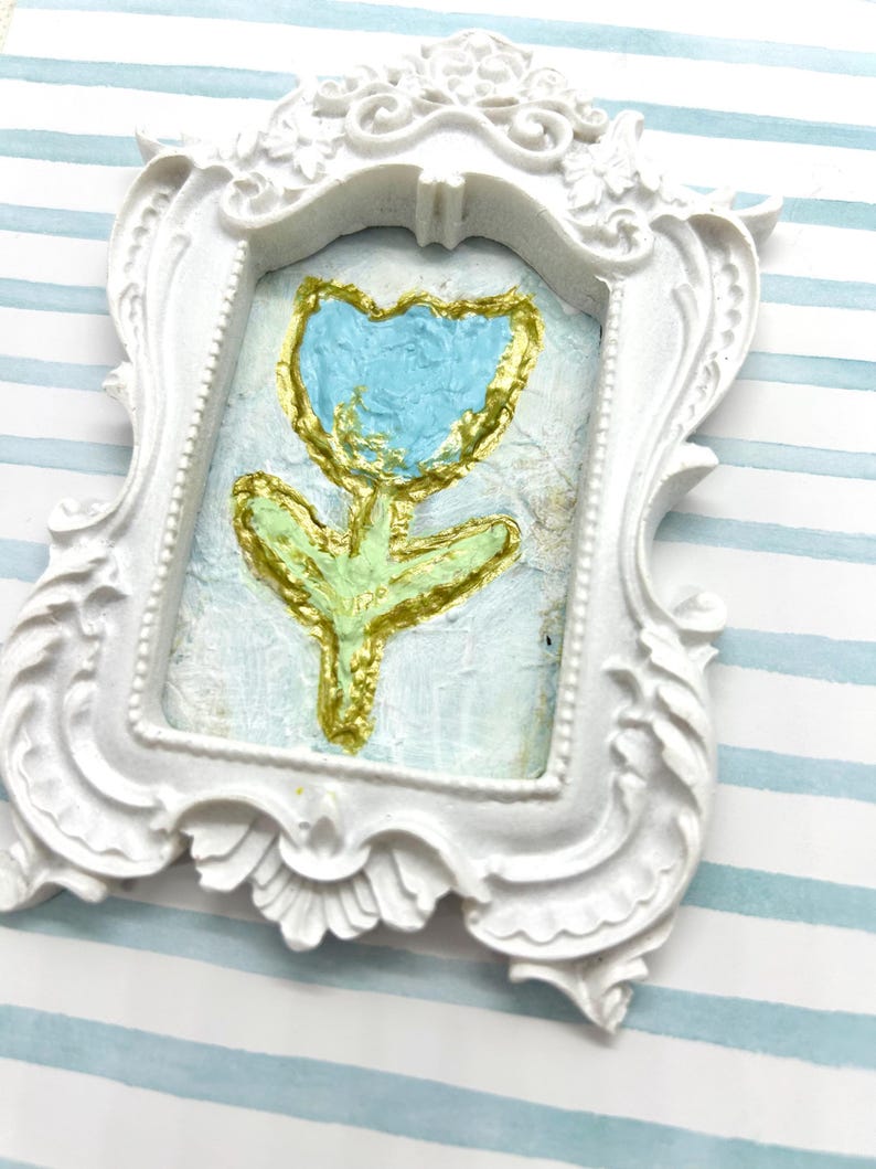 May include: A small, white, ornate picture frame with a painted tulip. The tulip has a blue flower, gold outline, and green stem and leaves. The background is a light blue color.