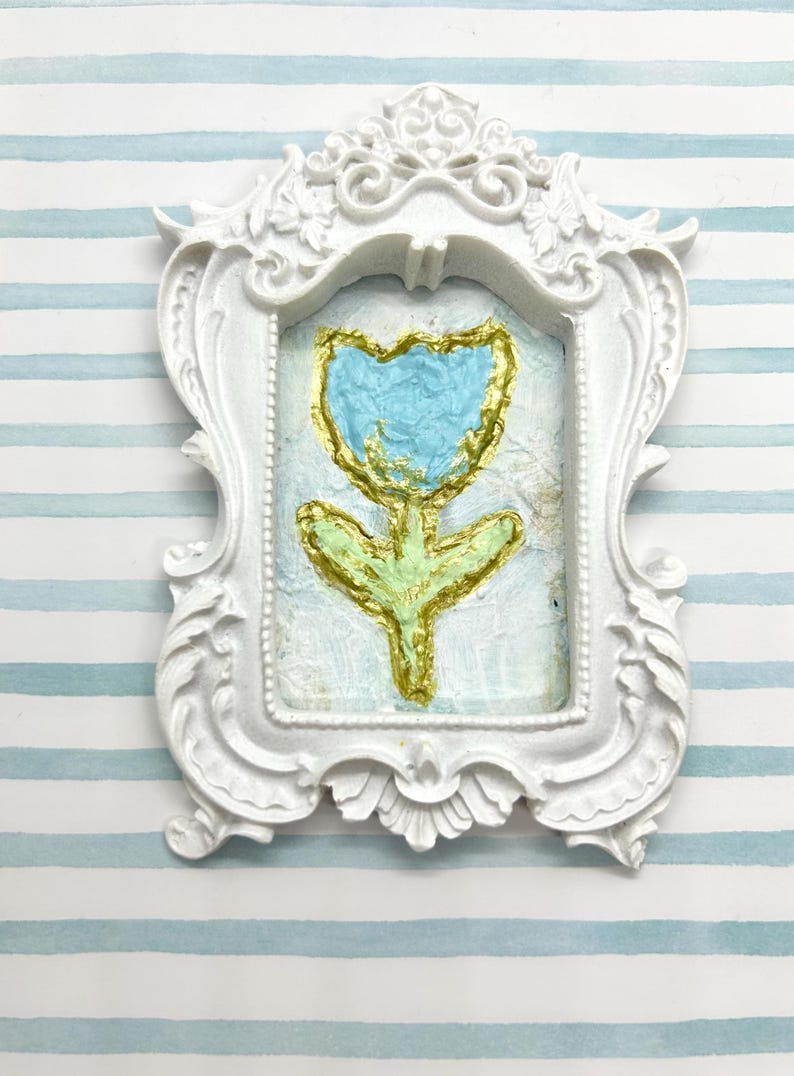 May include: A white, ornate picture frame with a painted flower. The flower has a light blue petal with gold outlining and a green stem. The background is a light blue and white striped pattern.