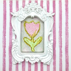 May include: A white ornate frame surrounds a painting of a pink tulip with gold accents and green leaves. The artwork is set against a pink and white striped background, creating a charming, decorative piece.