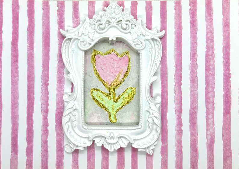 May include: A white ornate frame surrounds a painting of a pink tulip with gold accents and green leaves. The artwork is set against a pink and white striped background, creating a charming, decorative piece.