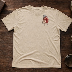 May include: A beige short-sleeved t-shirt featuring a small graphic on the upper left chest of a bird wearing a gray knit hat and a red scarf. The bird is depicted in a realistic style with brown and white feathers and orange accents.