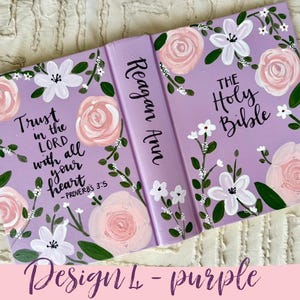 May include: A lilac Bible cover adorned with painted flowers. The design includes pink roses, white blossoms, and green foliage. The cover displays the text "Trust in the Lord with all your heart" and "The Holy Bible". The spine reads "Reagan Ann".