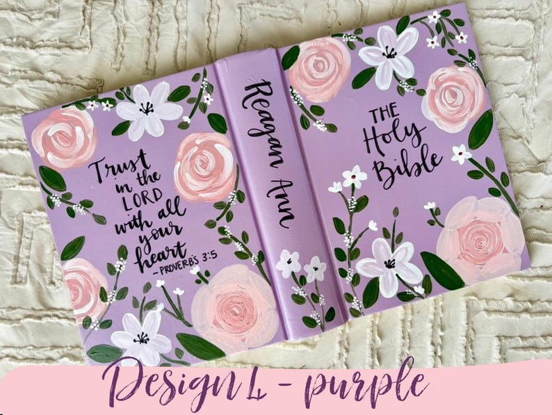 May include: A lilac Bible cover adorned with painted flowers. The design includes pink roses, white blossoms, and green foliage. The cover displays the text "Trust in the Lord with all your heart" and "The Holy Bible". The spine reads "Reagan Ann".