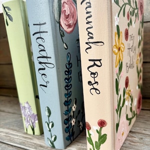 May include: Three decorative books with floral designs. The books are painted in pastel colours: light green, blue, and beige. The book spines have names written in black script: "Heather" and "Hannah Rose". The third book has the title "The Holy Bible".