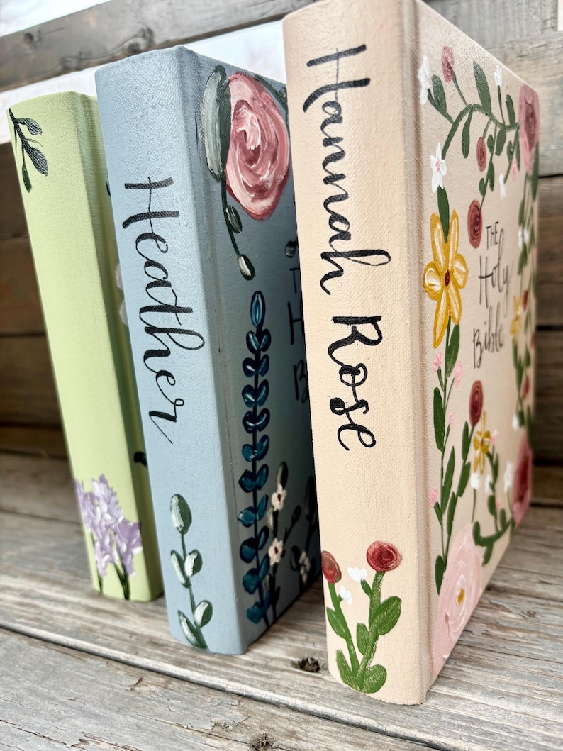 May include: Three decorative books with floral designs. The books are painted in pastel colours: light green, blue, and beige. The book spines have names written in black script: "Heather" and "Hannah Rose". The third book has the title "The Holy Bible".