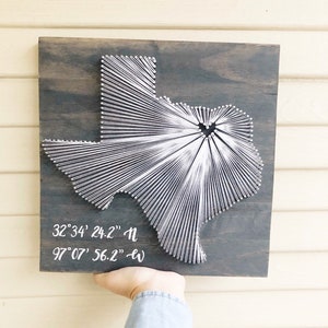May include: A square dark wood plaque featuring a white string art map of Texas. The string design radiates from a central point within the state shape. White text printed on the lower left reads 32°34' 24.2" N and 97°07' 56.2" W.