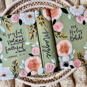 May include: A sage green Bible cover decorated with hand-painted flowers in pink, white, and peach. The cover features the text "Be joyful in hope, patient in affliction, faithful in prayer - Romans 12:12" and "The Holy Bible". The spine is personalized with the name "Maggie".