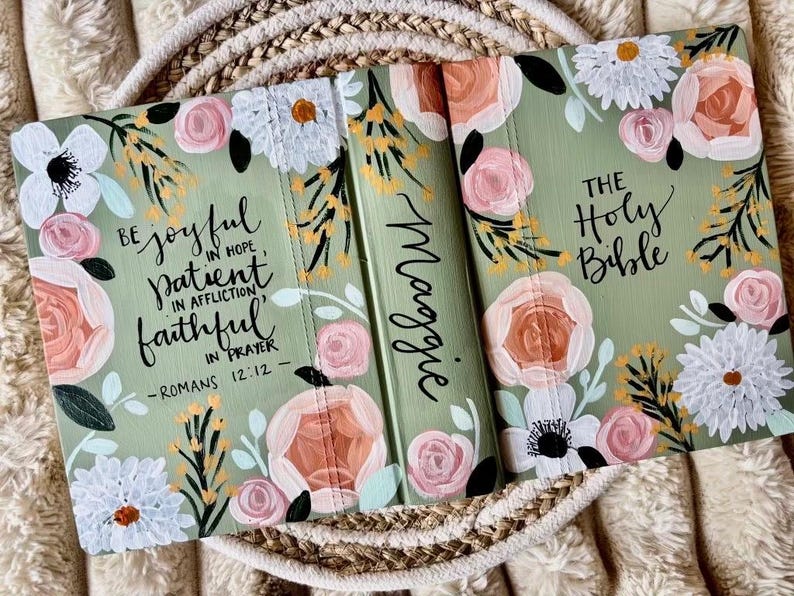 May include: A sage green Bible cover decorated with hand-painted flowers in pink, white, and peach. The cover features the text "Be joyful in hope, patient in affliction, faithful in prayer - Romans 12:12" and "The Holy Bible". The spine is personalized with the name "Maggie".
