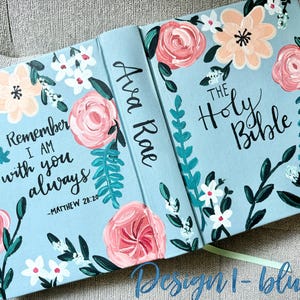May include: A light blue Bible cover with floral artwork. The cover features pink, peach, and white flowers with green leaves. Text includes "Remember I am with you always" and "The Holy Bible."