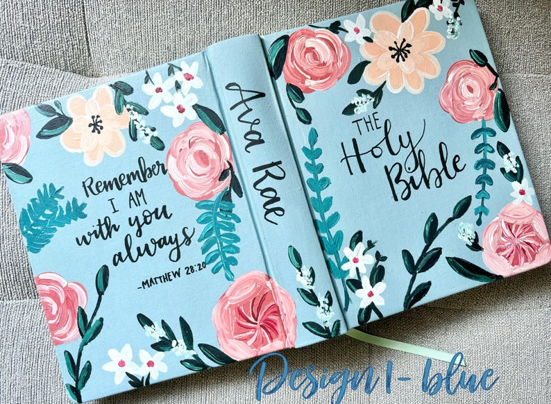 May include: A light blue Bible cover with floral artwork. The cover features pink, peach, and white flowers with green leaves. Text includes "Remember I am with you always" and "The Holy Bible."