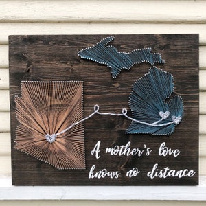 May include: Wooden string art featuring the states of Arizona and Michigan, connected by a string. The Arizona outline is filled with tan string, while Michigan is filled with blue string. The phrase "A mother's love knows no distance" is written in white.