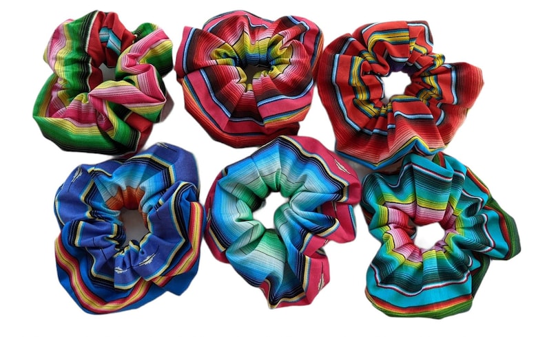 May include: Six colorful hair scrunchies with a Mexican serape pattern. The scrunchies are made of fabric and have a gathered look. The colors include red, green, blue, yellow, and white.