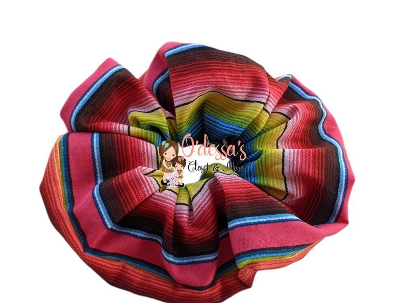 May include: A colorful hair scrunchie with a striped pattern reminiscent of a Mexican blanket. The scrunchie is made of fabric and is a bright pink, red, yellow, blue, and green.