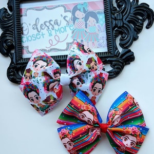 May include: Two colorful hair bows featuring a repeating pattern of Frida Kahlo's portrait. The bows are made of ribbon and have a white center.