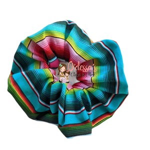 May include: A turquoise, green, yellow, red, and black striped scrunchie. The scrunchie is made of fabric and has a  "O'dessa's Closet & More" logo.