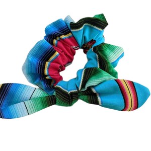 Puede incluir: Una colorida scrunchie con un vibrante patr&oacute;n de serape. La tela presenta rayas horizontales en tonos de azul, rosa, verde, amarillo y negro. La scrunchie est&aacute; dise&ntilde;ada con un detalle de lazo, agregando un toque decorativo al accesorio para el cabello.