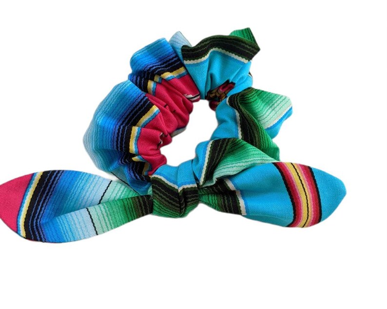 Puede incluir: Una colorida scrunchie con un vibrante patr&oacute;n de serape. La tela presenta rayas horizontales en tonos de azul, rosa, verde, amarillo y negro. La scrunchie est&aacute; dise&ntilde;ada con un detalle de lazo, agregando un toque decorativo al accesorio para el cabello.
