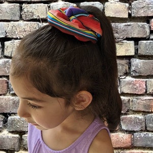 May include: A young girl with long brown hair wearing a purple tank top and a colorful striped hair scrunchie.