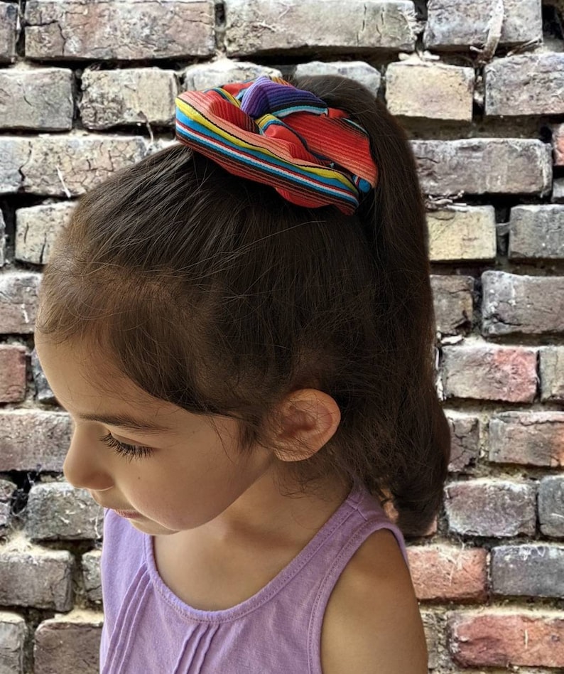 May include: A young girl with long brown hair wearing a purple tank top and a colorful striped hair scrunchie.