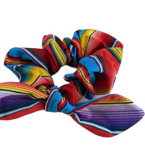 Puede incluir: Una colorida scrunchie con lazo, con un vibrante patr&oacute;n de rayas en rojo, azul, amarillo y morado. La tela est&aacute; fruncida y atada en forma de lazo, creando un elegante accesorio para el cabello.