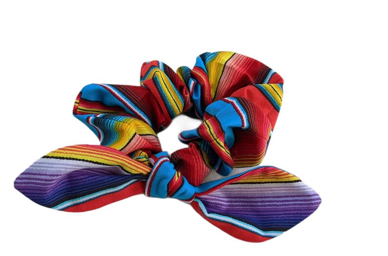 Puede incluir: Una colorida scrunchie con lazo, con un vibrante patr&oacute;n de rayas en rojo, azul, amarillo y morado. La tela est&aacute; fruncida y atada en forma de lazo, creando un elegante accesorio para el cabello.
