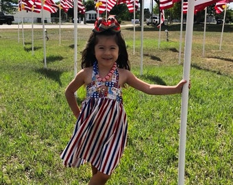 May include: A young girl wearing a red, white, and blue striped dress and a patriotic bow in her hair. She is standing in a grassy field with numerous American flags in the background, smiling at the camera.
