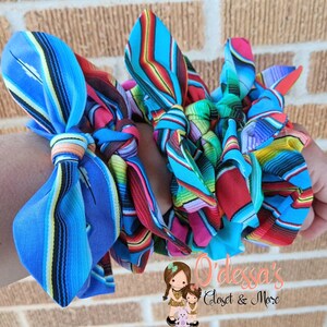 Puede incluir: Una colecci&oacute;n de coloridas gomas para el pelo de tela con un vibrante estampado de rayas en tonos azules, rojos, verdes y amarillos. Las gomas para el pelo est&aacute;n anudadas y parecen hechas a mano. La imagen incluye el texto "O'dessa's Closet & More".