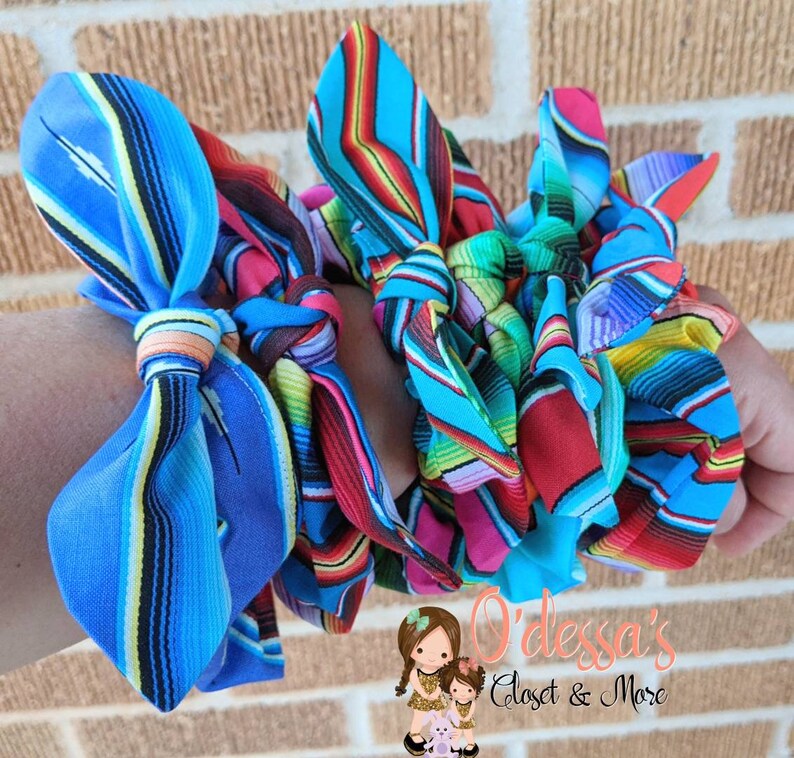Puede incluir: Una colecci&oacute;n de coloridas gomas para el pelo de tela con un vibrante estampado de rayas en tonos azules, rojos, verdes y amarillos. Las gomas para el pelo est&aacute;n anudadas y parecen hechas a mano. La imagen incluye el texto "O'dessa's Closet & More".