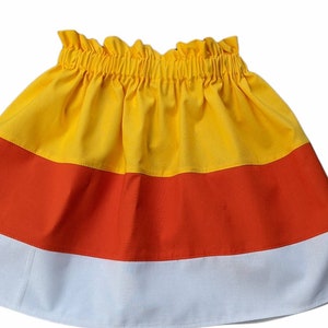 Pode incluir: Uma saia com tema de doces de milho, com um esquema de cores amarelo, laranja e branco. A saia tem uma cintura franzida e uma silhueta rodada. A pe&ccedil;a de roupa &eacute; feita de tecido e provavelmente &eacute; para uma crian&ccedil;a.