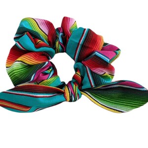 Puede incluir: Una colorida scrunchie con un vibrante patr&oacute;n de serape. La tela presenta rayas horizontales en turquesa, rojo, rosa, amarillo y verde. La scrunchie tiene un detalle de lazo, agregando un toque decorativo.