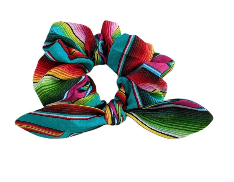 Puede incluir: Una colorida scrunchie con un vibrante patr&oacute;n de serape. La tela presenta rayas horizontales en turquesa, rojo, rosa, amarillo y verde. La scrunchie tiene un detalle de lazo, agregando un toque decorativo.