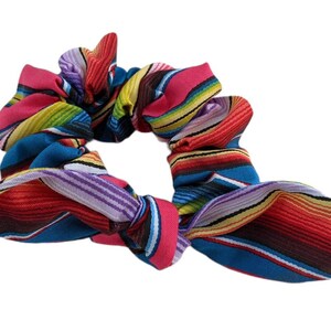 Puede incluir: Una colorida scrunchie con un vibrante estampado de rayas en tonos rojos, rosas, azules, amarillos y morados. La tela est&aacute; fruncida y atada en forma de lazo, perfecta para a&ntilde;adir un toque de color a cualquier peinado.