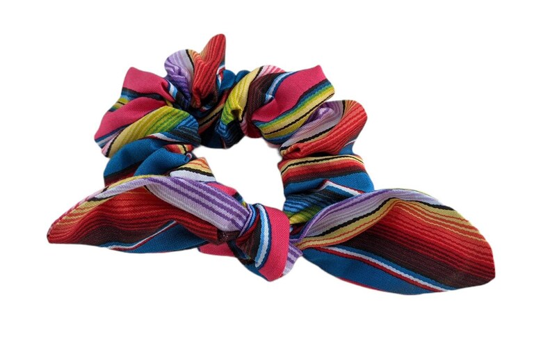 Puede incluir: Una colorida scrunchie con un vibrante estampado de rayas en tonos rojos, rosas, azules, amarillos y morados. La tela est&aacute; fruncida y atada en forma de lazo, perfecta para a&ntilde;adir un toque de color a cualquier peinado.