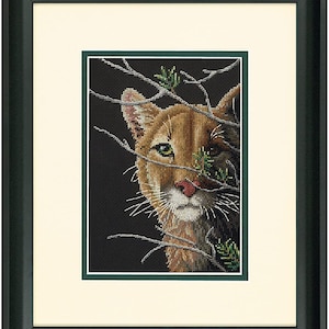 May include: Framed cross-stitch art featuring a cougar peeking through branches. The cougar has tan fur, a pink nose, and green eyes. The artwork is framed in a black frame with a white mat and a green inner border. The background is black.
