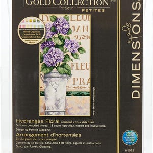 May include: Hydrangea Floral counted cross stitch kit by Dimensions. The kit includes presorted thread, 18 count ivory Aida fabric, a needle, and instructions. The design features a bouquet of purple hydrangeas in a white metal container with a crown on the side. The design is by Pamela Gladding. The kit measures 4 inches by 8 inches.