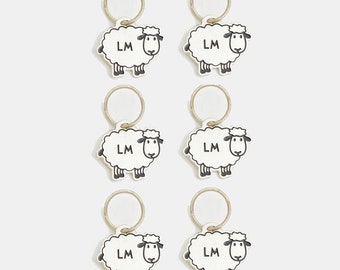 May include: Six white sheep charms with black eyes and a black nose. Each charm has a gold ring and the letters "LM" printed on the sheep's body.