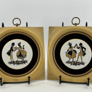 May include: Two square wall plaques featuring circular white ceramic centers with black and gold silhouettes of figures in period clothing. Each plaque has a textured gold frame, a black circular border, and a brass hanging ring at the top.